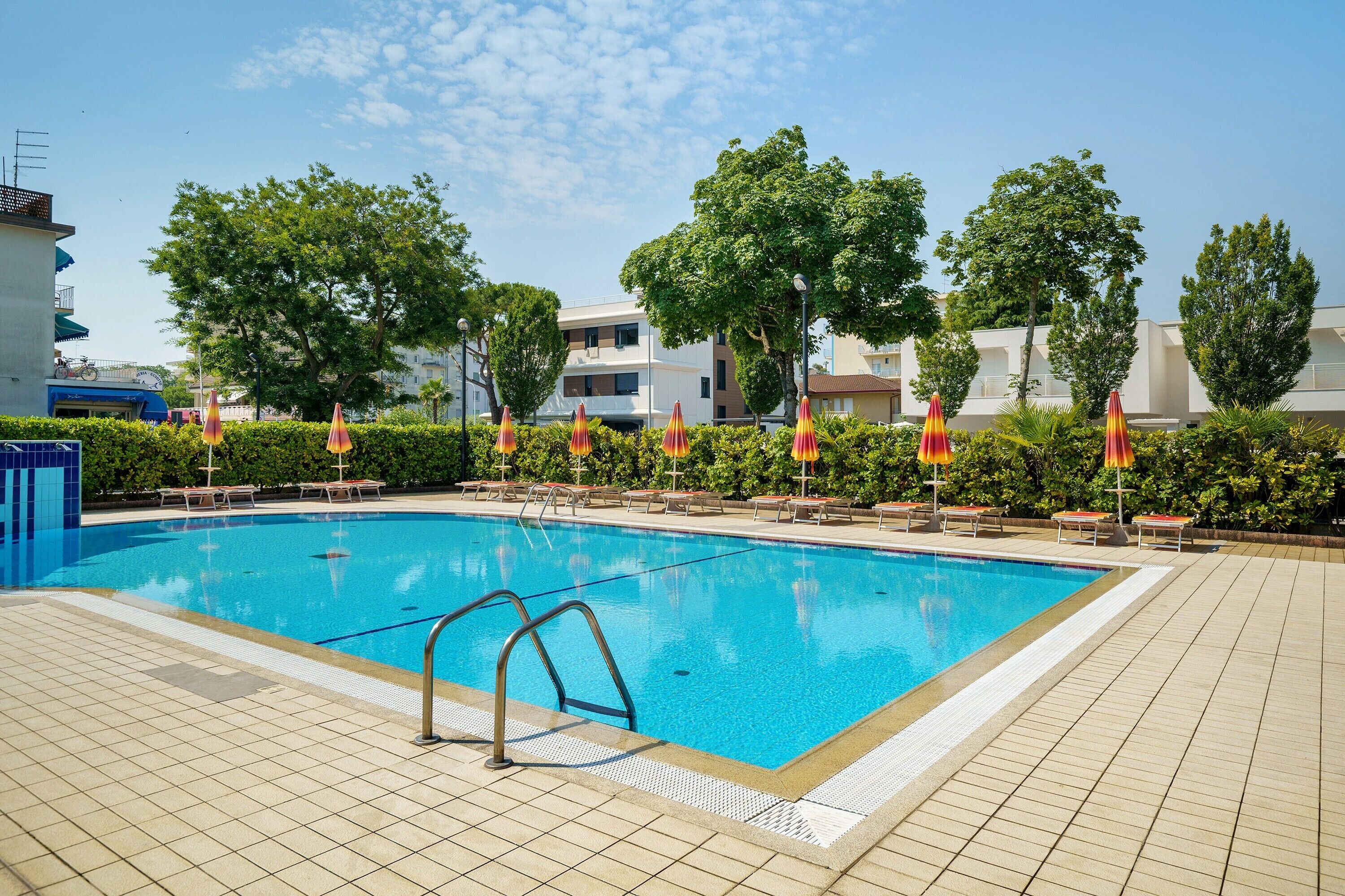 A swimming pool with sun loungers and umbrellas, surrounded by trees and buildings.
