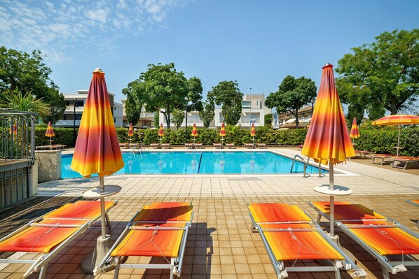 Ein Schwimmbecken mit Liegestühlen und Sonnenschirmen, umgeben von Bäumen und Gebäuden.