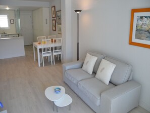 A modern living room with a sofa, two small tables, a dining area with chairs, a kitchen area, and a framed picture on the wall.