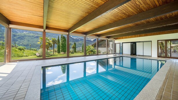 A swimming pool with a wooden ceiling and large windows overlooking a mountainous landscape.