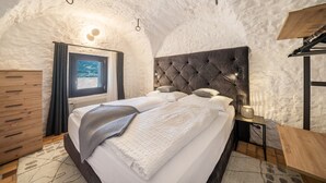A bedroom with a tufted headboard, a bed with white linens, a wooden nightstand, a wall-mounted lamp, and a window with curtains.