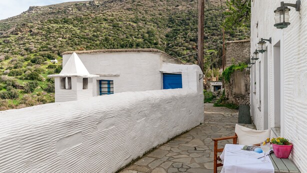 A white building with a blue door and window, a stone pathway, and a table with a chair and a potted plant. - Olive Guesthouse Junior Apartment with Sea View and Wi-Fi (Andros island)