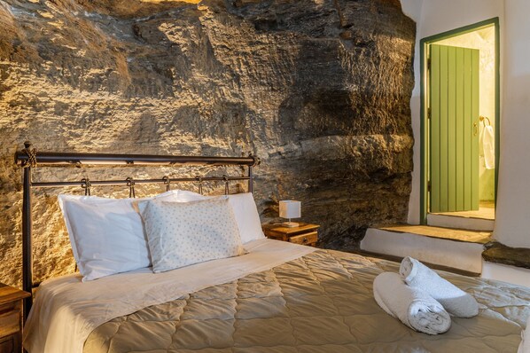 Una cama con cubrecama acolchado, almohadas y una toalla enrollada en una habitación con pared de piedra y puerta verde.