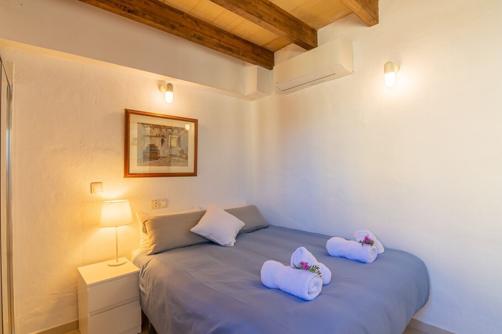 Un dormitorio con cama, mesita de noche, lámpara, un cuadro enmarcado y dos toallas sobre la cama.