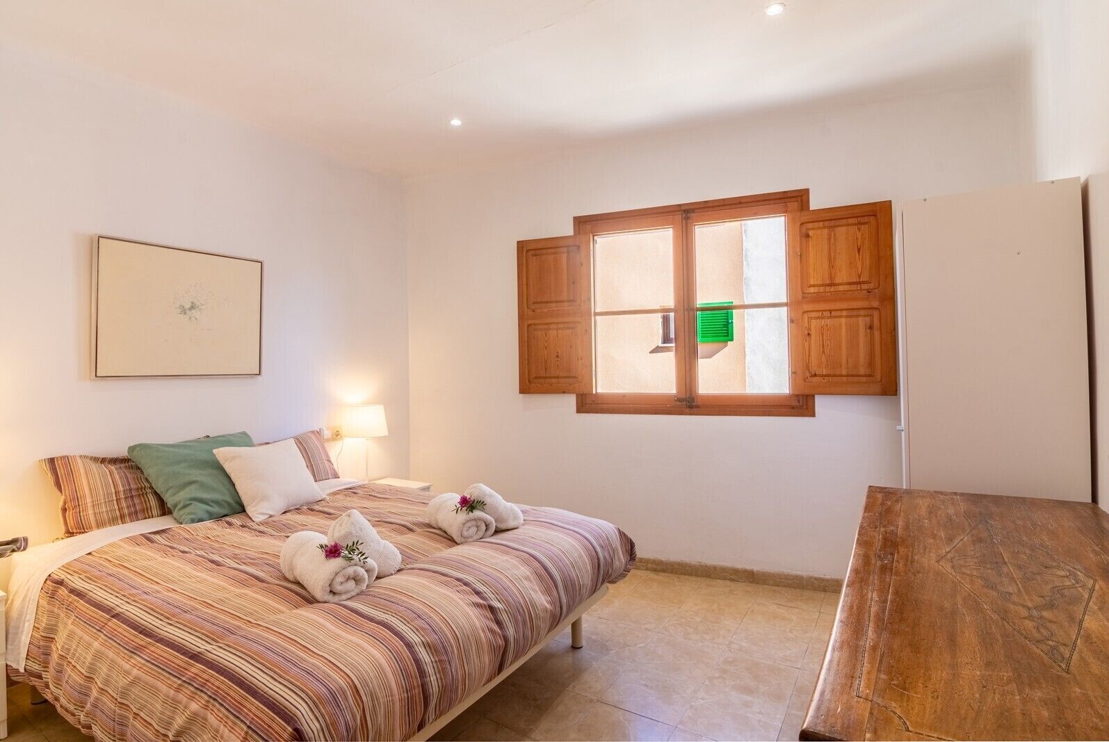 A bedroom with a bed, pillows, a bedside table, a window with wooden shutters, and a framed picture on the wall.