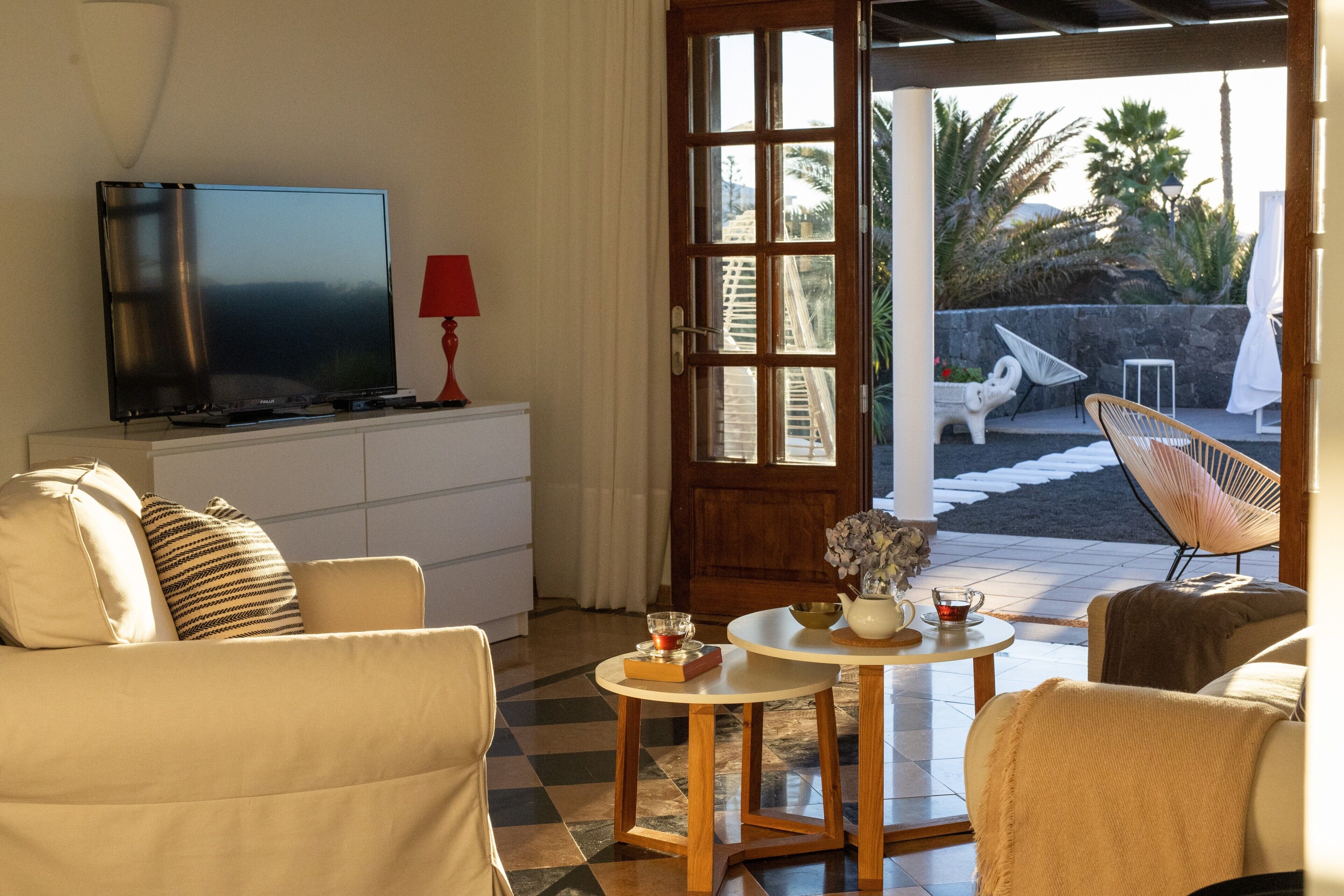 Uma sala de estar com televisão, sofá, cadeiras, uma mesinha de centro com um bule e vista para o pátio.