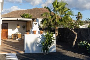 Une maison avec un patio, un mur blanc, une porte en bois, une table avec des chaises, et une montagne en arrière-plan.