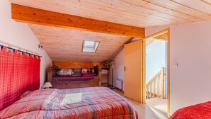 Une chambre avec un plafond en pente, un puits de lumière, un lit recouvert d’une couvre-lit à motifs rouges et blancs, une tête de lit en bois et un rideau rouge.