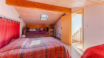 Une chambre avec un plafond en pente, un puits de lumière, un lit recouvert d’une couvre-lit à motifs rouges et blancs, une tête de lit en bois et un rideau rouge.