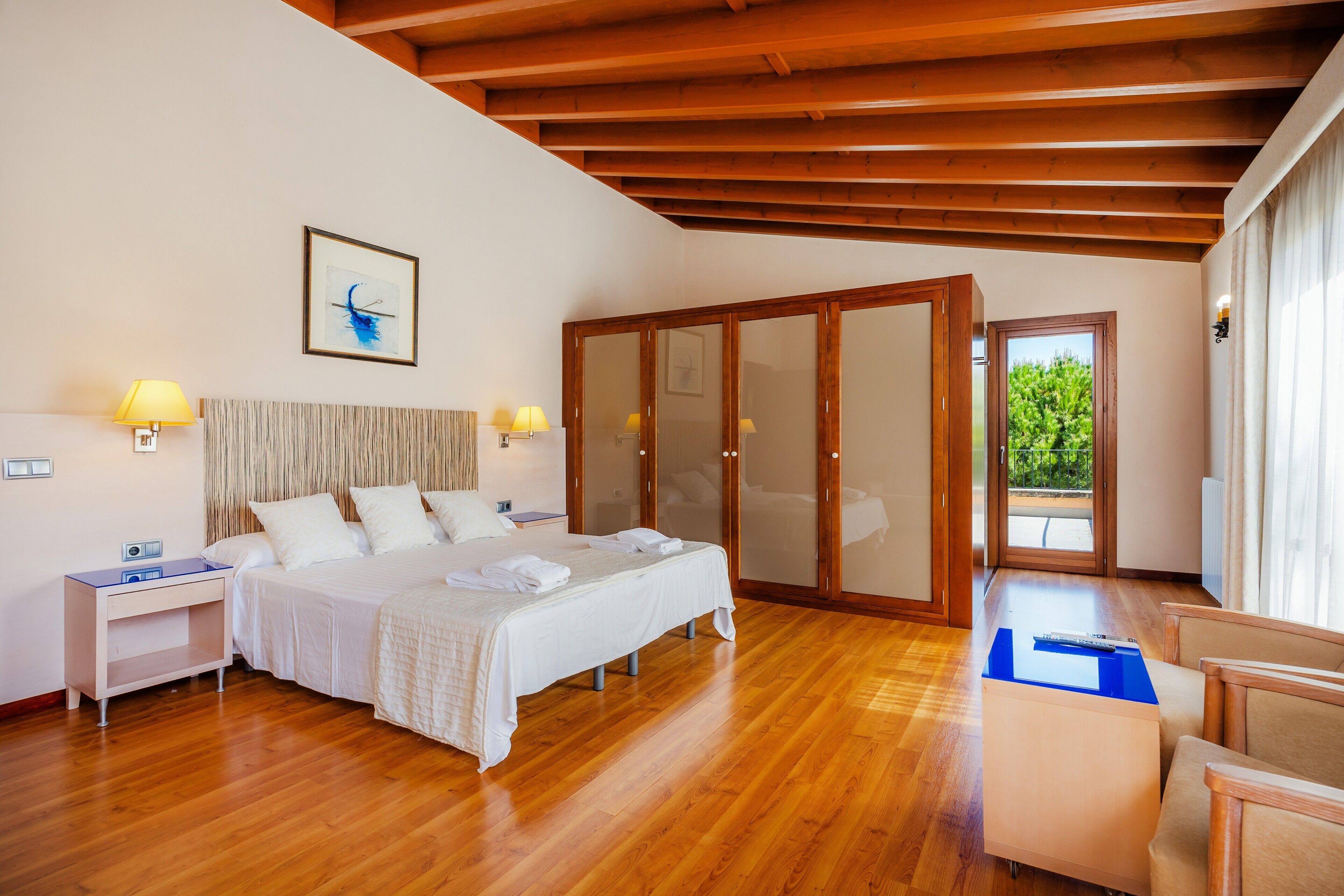 A hotel room with a bed, bedside tables, a desk, a chair, a wardrobe, and a view of greenery outside.