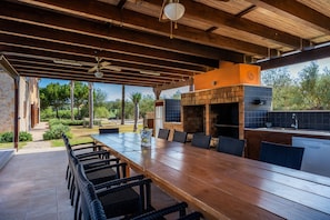 An outdoor patio with a long wooden table and chairs, a stone wall, and a brick fireplace.