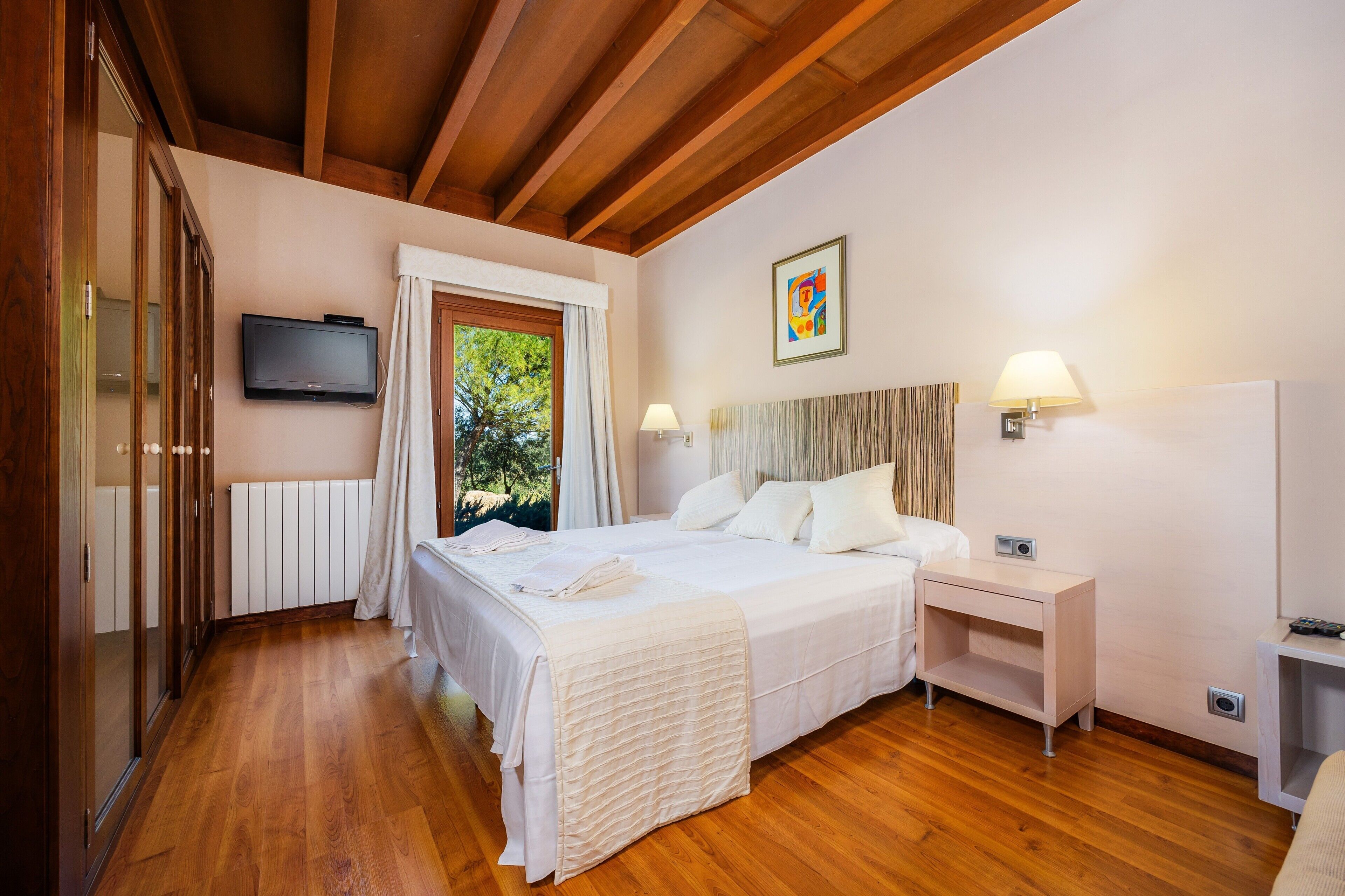 A bedroom with a wooden floor, a bed with white linens, a bedside table with a lamp, and a TV mounted on the wall.