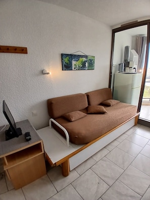 A modern room with a brown sofa, a small wooden table with a monitor and remote, a wall-mounted picture, and a sliding glass door leading to a balcony.
