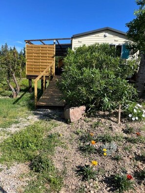 A wooden structure with a ramp leading to a house, surrounded by greenery and flowering plants.
