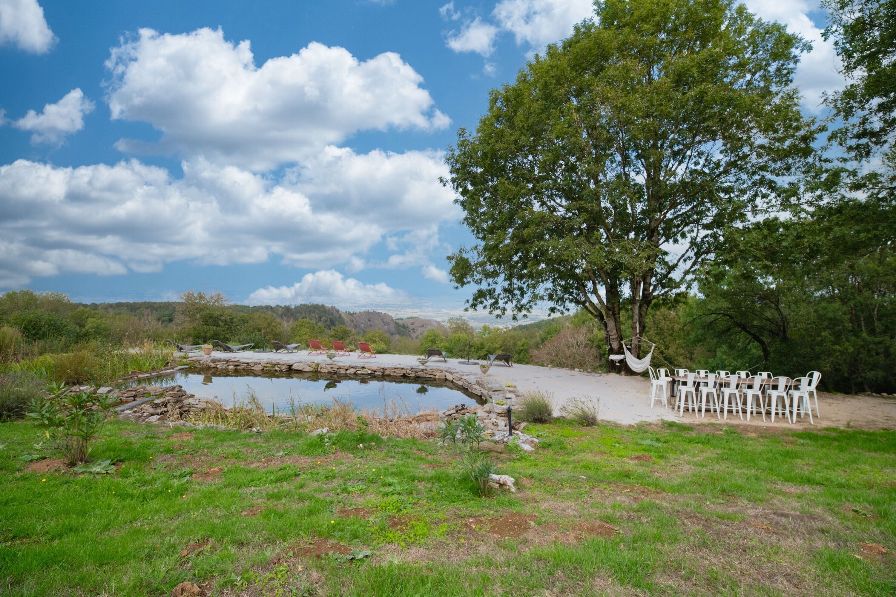 A natural outdoor setting with a pond, a large tree, and a seating area with chairs.