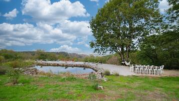 A natural outdoor setting with a pond, a large tree, and a seating area with chairs.