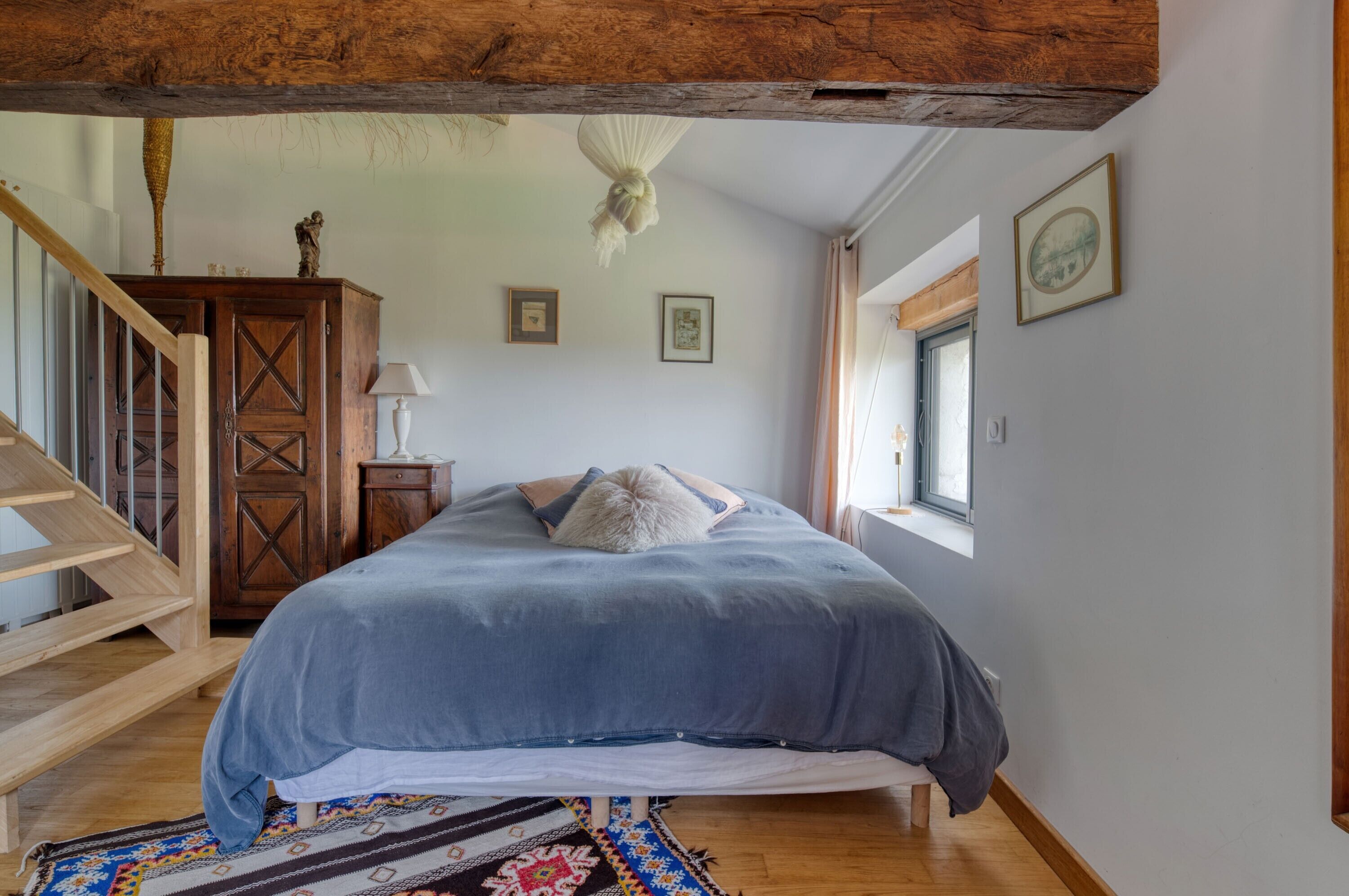 A bedroom with a bed, wooden floor, a staircase, a wooden cabinet, a lamp, and framed pictures on the wall.