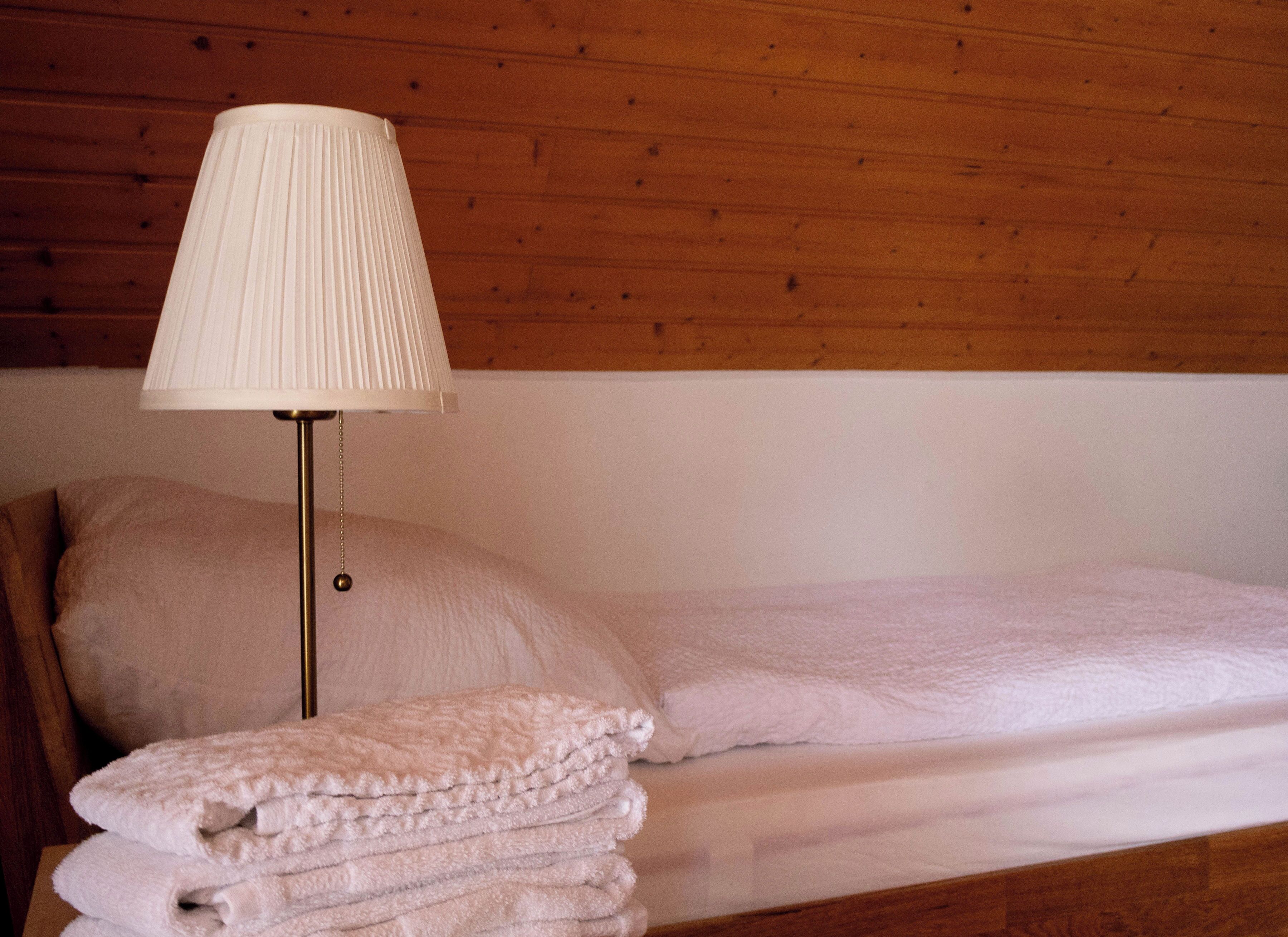 A neatly made bed with a white comforter and pillows, a lamp with a white shade, and a stack of folded towels.