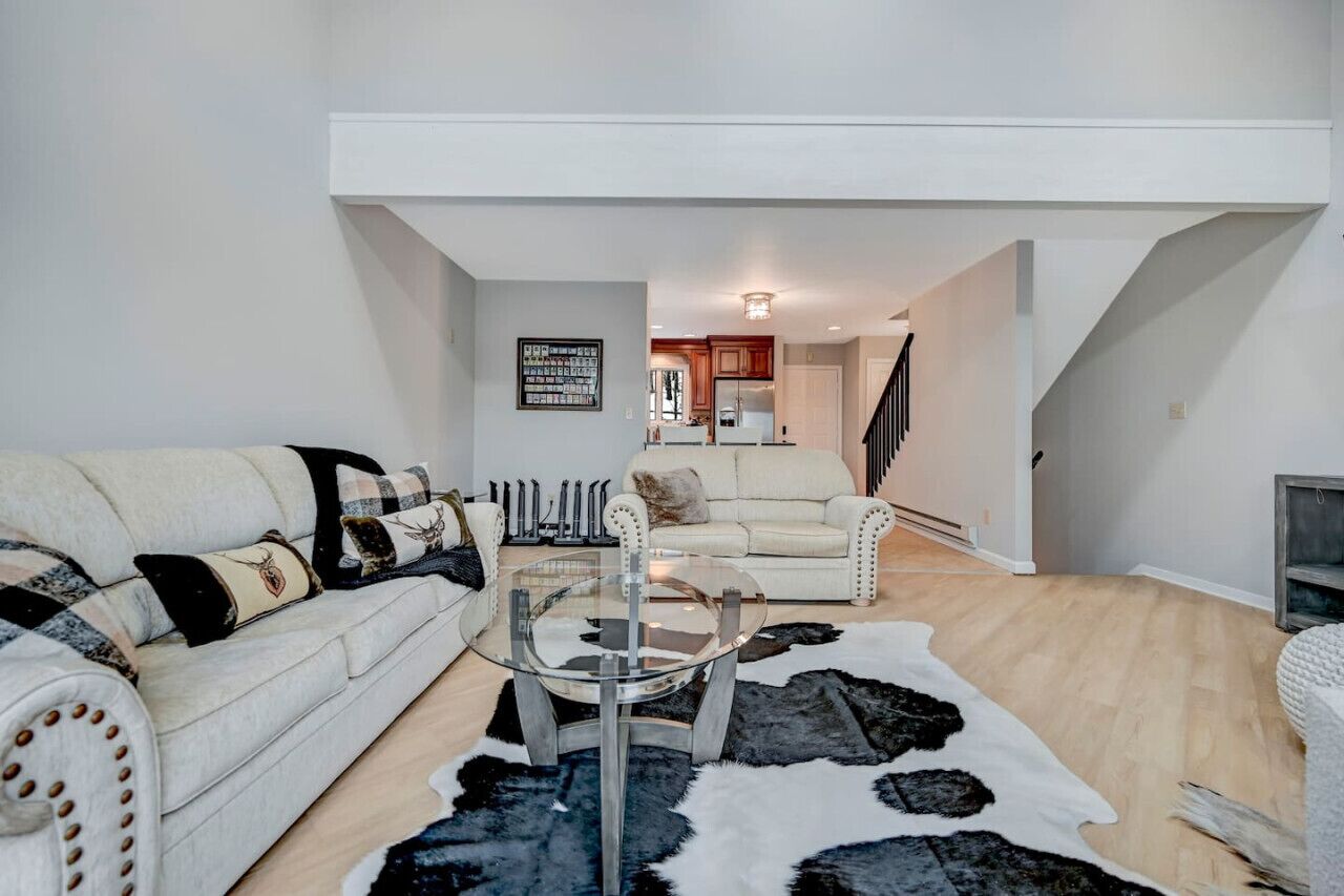 A modern living room with a glass coffee table, two sofas, a black and white rug, and a kitchen in the background.