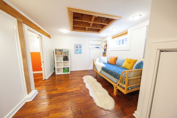 Una habitación con suelo de madera, un tapete blanco y mullido, un sofá de mimbre con cojines de colores, un techo de madera con vigas a la vista y una puerta blanca.