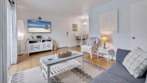 Een woonkamer met een televisie, witte meubels, een kamerbreed tapijt met dessin en een muur met een schilderij van een zeilboot.