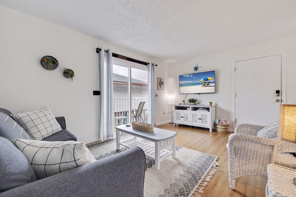A living room with a grey sofa, a white coffee table, wicker chairs, a TV on the wall, and a view of the beach through the window.