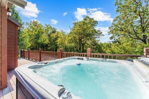 A hot tub with a wooden deck and a metal railing, surrounded by trees and a clear sky.