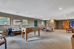 Uma área de lazer ampla e coberta, com várias mesas de futebol de mesa e uma mesa de bilhar, paredes de madeira e grandes janelas.