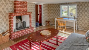 Una sala de estar con chimenea de ladrillo, una mesa redonda de madera, un tapete a rayas rojas y blancas, y un ventanal con vistas al exterior.