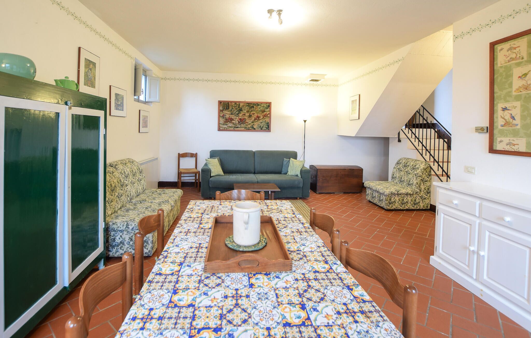 A cozy living room with a green sofa, a dining table with a colorful tablecloth, and a staircase leading to another level.