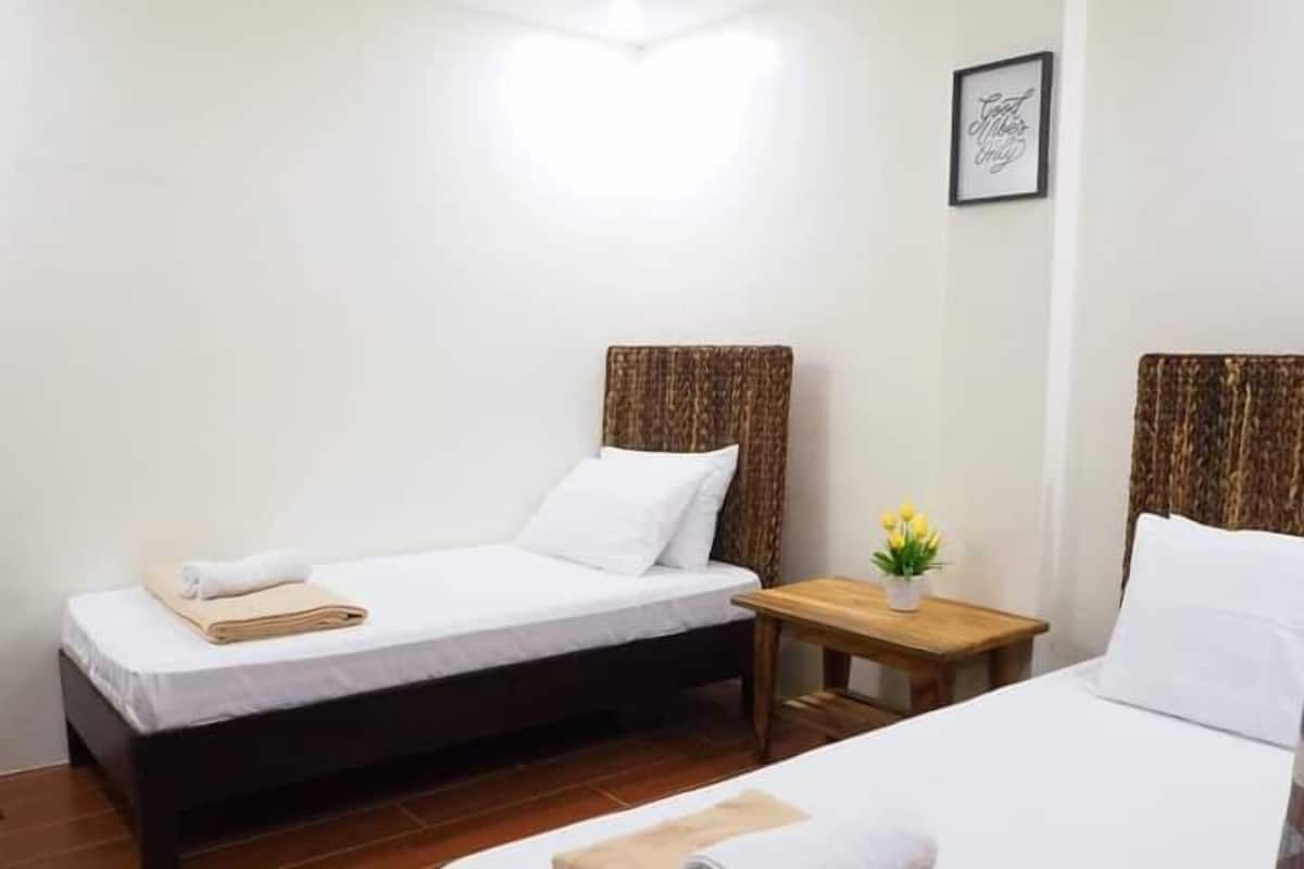 A hotel room with two single beds, each with a wooden headboard, white bedding, and a rolled towel on the bed. There is a small wooden table with a plant between the beds.