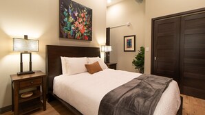 A neatly made bed with white linens and a gray throw, a wooden nightstand with a lamp, a painting above the bed, and a closed door to the right.