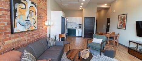 A modern living room with a sofa, coffee table, and chairs. There is a kitchen in the background with white cabinets and a dishwasher.
