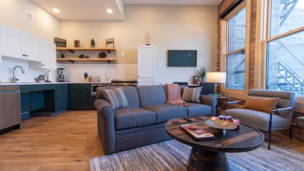 A modern kitchen with white cabinets, a built-in oven, and a coffee machine. There is a gray sofa with a striped rug in the living area, and a round coffee table with magazines and a remote control. - Luxury Craft District Suites | Unit 102 (Duluth)