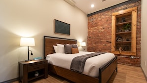 A modern bedroom with a wooden bed, two bedside tables, a brick accent wall, and a wooden shelf with decorative items. - Luxury Craft District Suites | Unit 102 (Duluth)