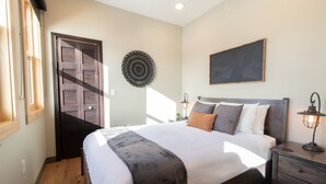 Une chambre avec un grand lit, une porte en bois, une assiette décorative accrochée au mur, une table de chevet avec une lampe et une fenêtre avec des rideaux.