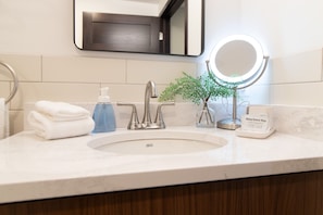Un baño con lavamanos, un espejo con luz, dispensador de jabón, una planta y dispensador de toallitas desmaquillantes.