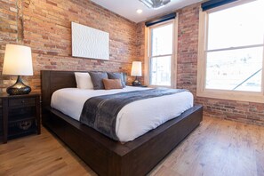 Quarto com parede de tijolos, uma janela grande, cama de madeira e duas luminárias de cabeceira.