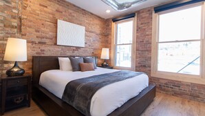 Un dormitorio con pared de ladrillo, una cama grande, dos mesitas de noche con lámparas y una ventana con vistas.