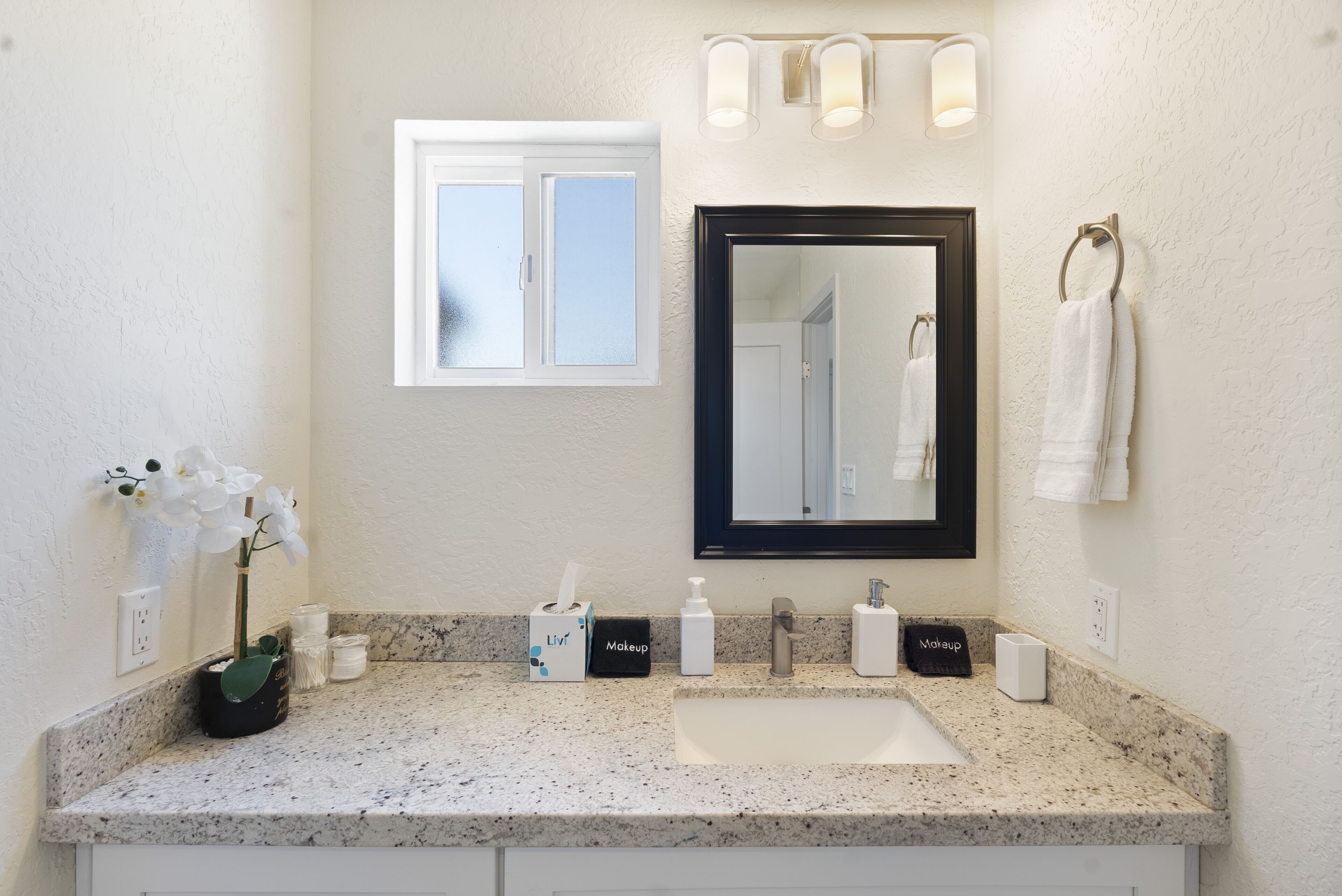 A bathroom with a granite countertop, a white sink, a black-framed mirror, and a window above the mirror.