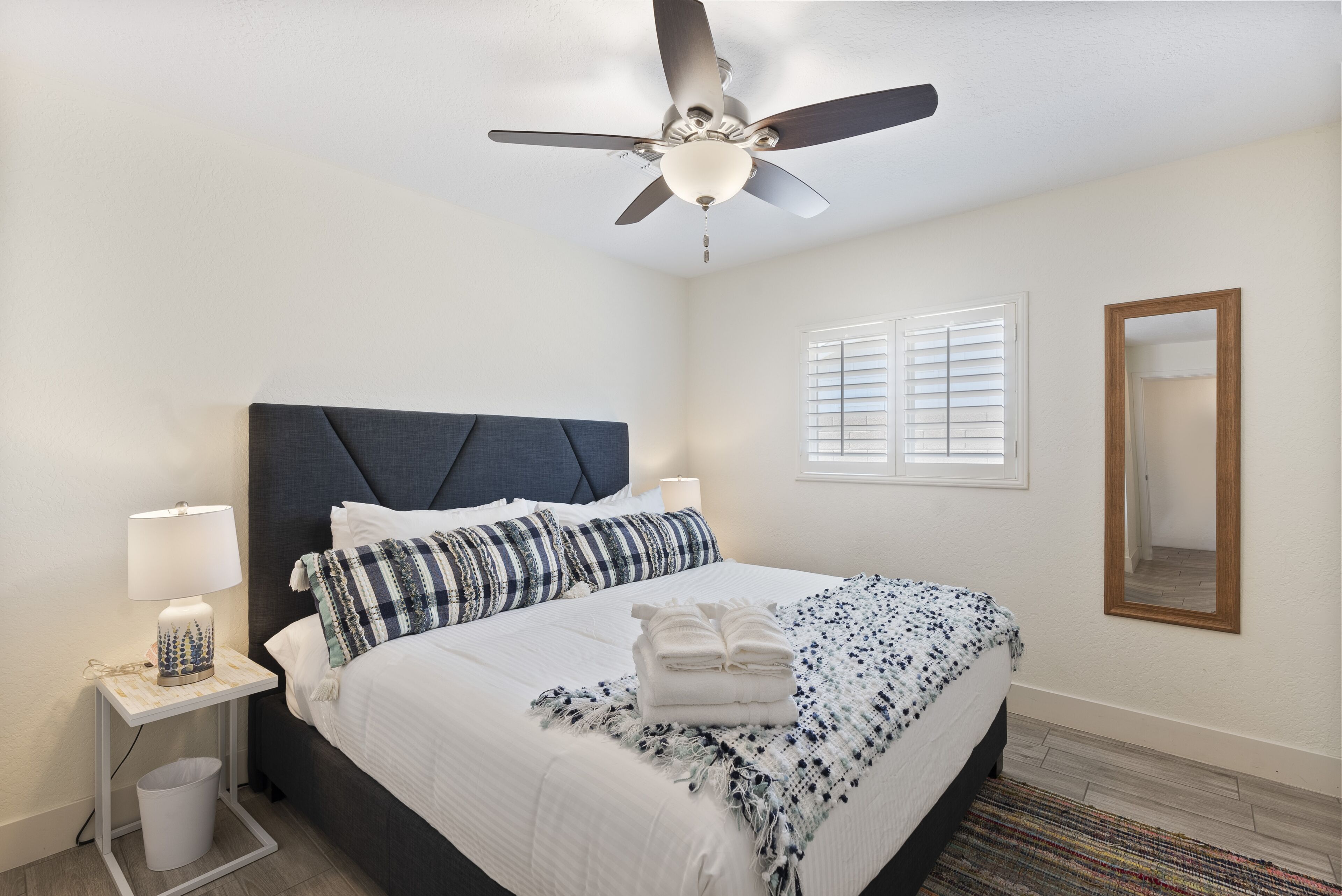 A neatly arranged bedroom with a bed, bedside lamps, a ceiling fan, a window with blinds, and a mirror on the wall.