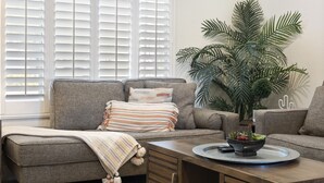 Un salon avec un canapé gris, un plaid à rayures, une table basse sur laquelle se trouve un saladier rempli de fruits, et une grande plante en pot.
