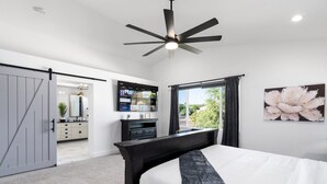 A bedroom with a bed, a ceiling fan, a TV, a window with curtains, and a door leading to another room. - Casa Yucca 1 (Chandler)