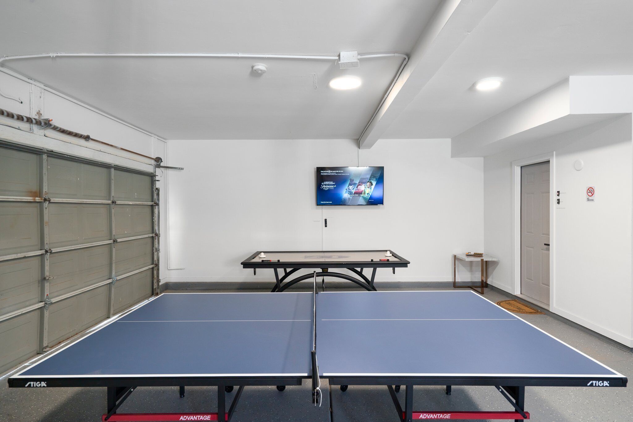 Een tafeltennistafel in een kamer met witte muren, een wandgemonteerde televisie en een gesloten garageklep.