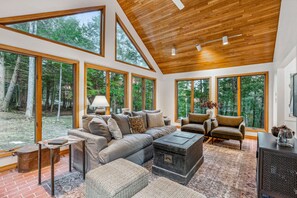 A spacious living room with a large sofa, armchairs, and a coffee table. There are large windows offering views of a forested area. - Twin Peaks Saltbox with Sauna, Pet-Friendly (Quechee)