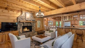 Interior de una cabaña de troncos con chimenea de piedra, sofás blancos, televisor y zona de cocina con armarios de madera y una barra.