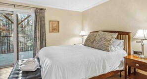 Une chambre avec un grand lit, une table de chevet avec une lampe, une tête de lit en bois, une fenêtre avec des rideaux et une vue sur les arbres à l’extérieur.