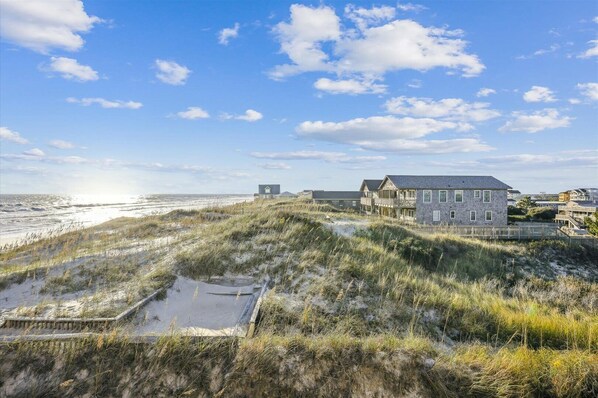 Une propriété située en bord de mer, comprenant une grande maison et un bâtiment plus petit, le tout entouré de dunes et d’une plage de sable.