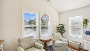 A cozy room with two large windows, a round wooden table, two striped armchairs, a blue patterned rug, and a potted plant.