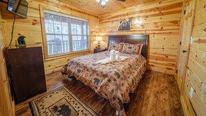 A wooden cabin bedroom with a bed, a ceiling fan, a window with blinds, a small desk, and a bear statue.